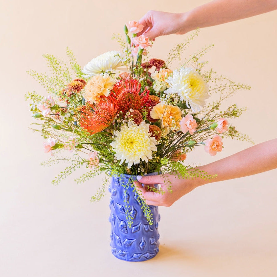 Manos arreglando el ramo de verano de Colvin La Jarana con crisantemos, nutans naranjas y flores crema en un jarrón azul con relieve.