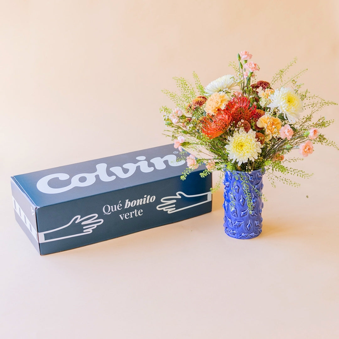 Ramo de flores La Jarana en un jarrón azul junto a la caja de regalo de Colvin con el mensaje "Qué bonito verte".