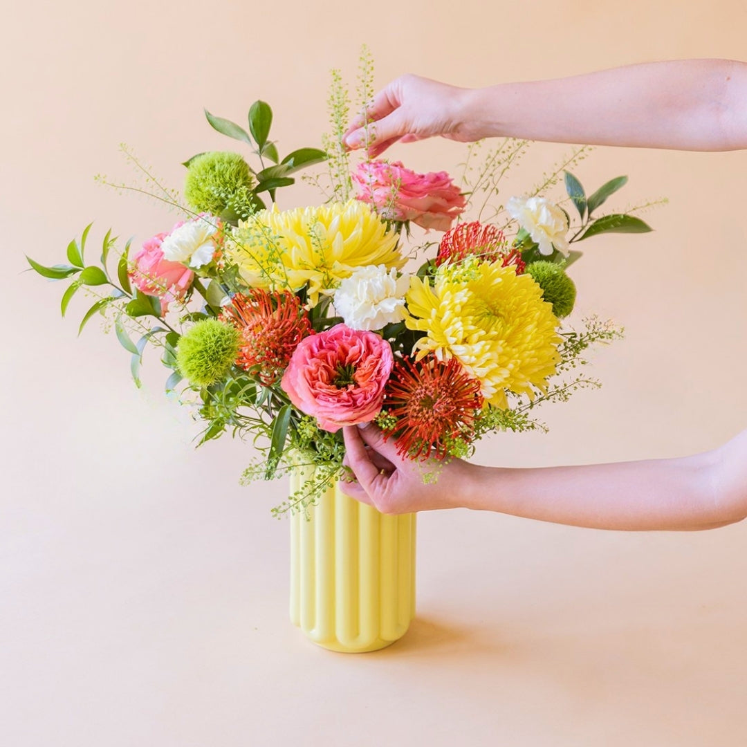 Manos arreglando el ramo de verano de Colvin La Verbena con crisantemos amarillos, nutans y rosas de jardín en un jarrón amarillo.