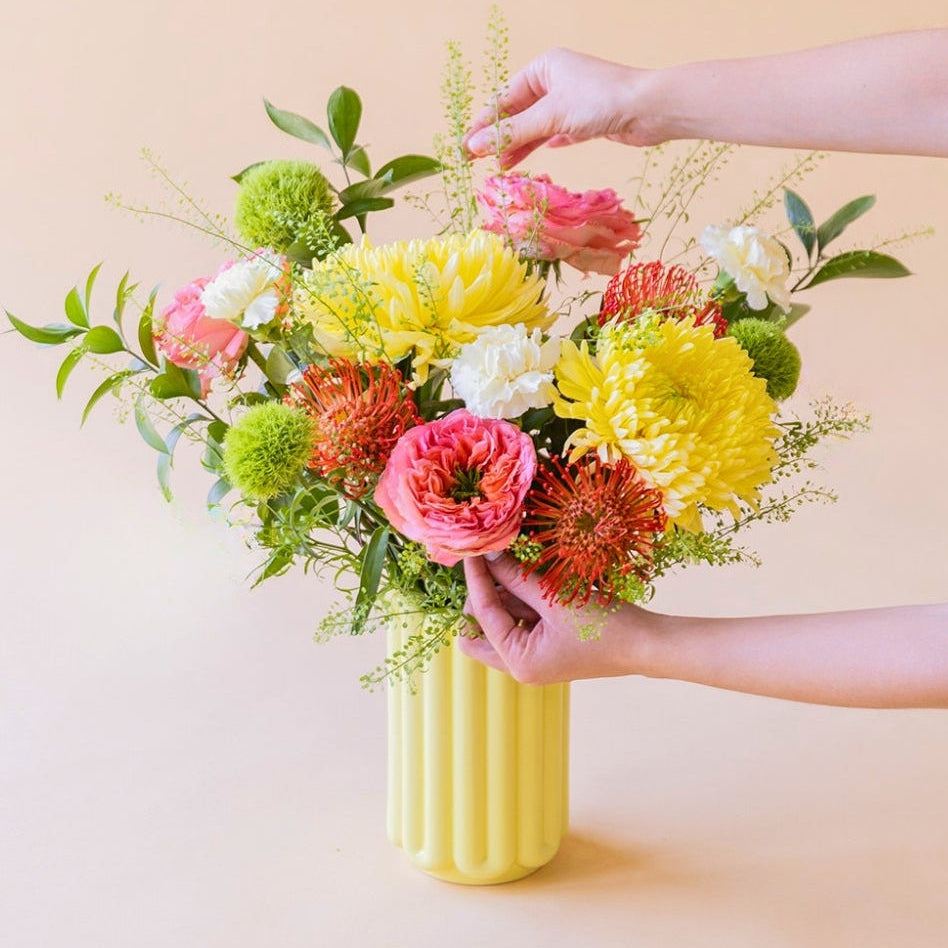 Manos arreglando el ramo de verano de Colvin La Verbena con crisantemos amarillos, nutans y rosas de jardín en un jarrón amarillo.
