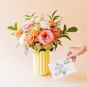 Ramo Colvin de rosas garden melocotón, achillea y aster crema en jarrón de cerámica Alba amarillo, junto a tarjeta ¡Hola peque!s en jarrón Alba amarillo, con mano sosteniendo tarjeta ¡Hola peque!