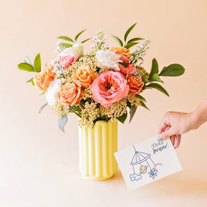 Ramo Colvin de rosas garden melocotón, achillea y aster crema en jarrón de cerámica Alba amarillo, junto a tarjeta ¡Hola peque!s en jarrón Alba amarillo, con mano sosteniendo tarjeta ¡Hola peque!