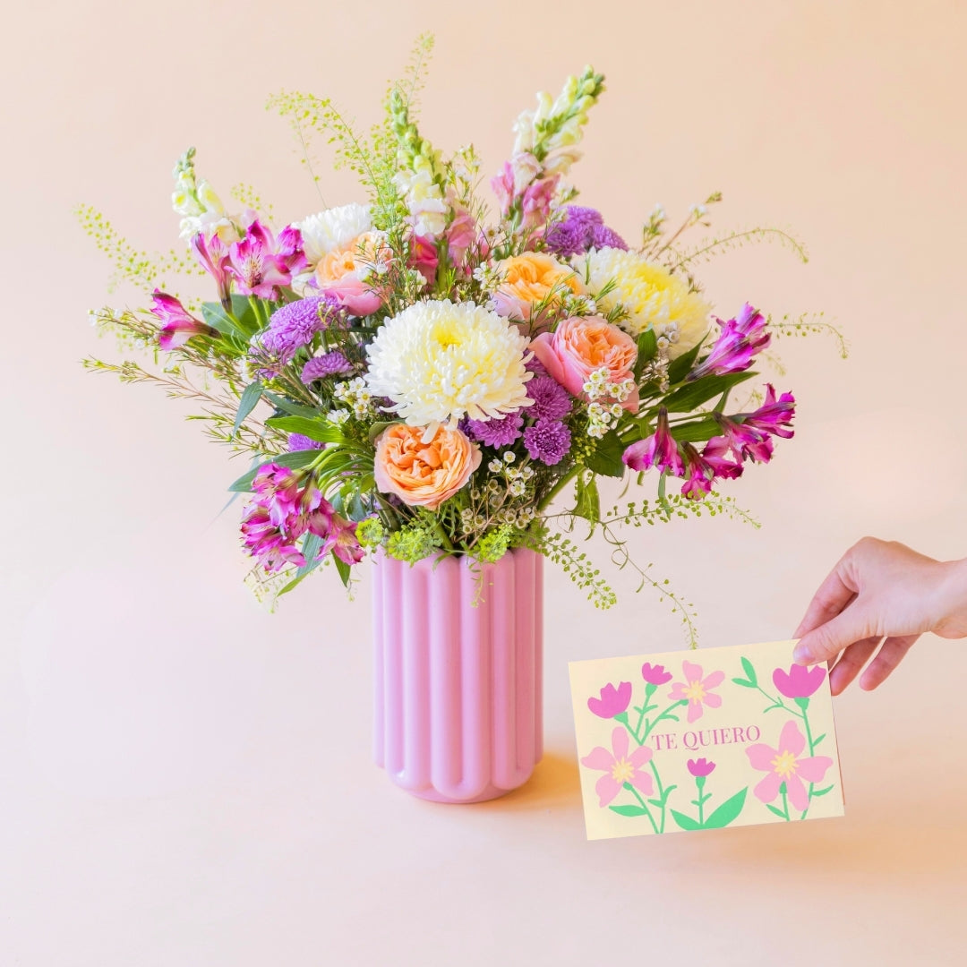 La Costa - Arreglo floral de verano en jarrón rosa con mano sosteniendo tarjeta "Te Quiero".