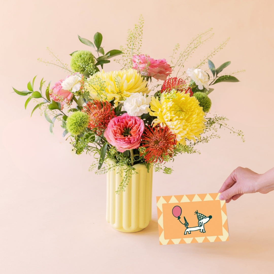 Ramo Verbena con flores de verano, rosas de jardín y nutans, con jarrón de cerámica amarillo y mano con tarjeta de cumpleaños con perrito y globo. 