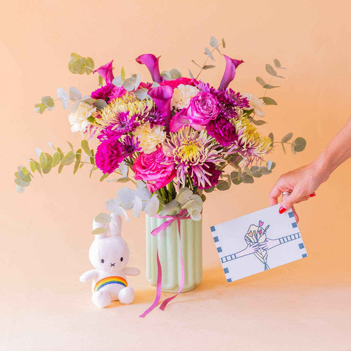 Mano sosteniendo la tarjeta Más que Flores junto al Ramo Euforia de calas moradas y peluche Miffy — Colvin