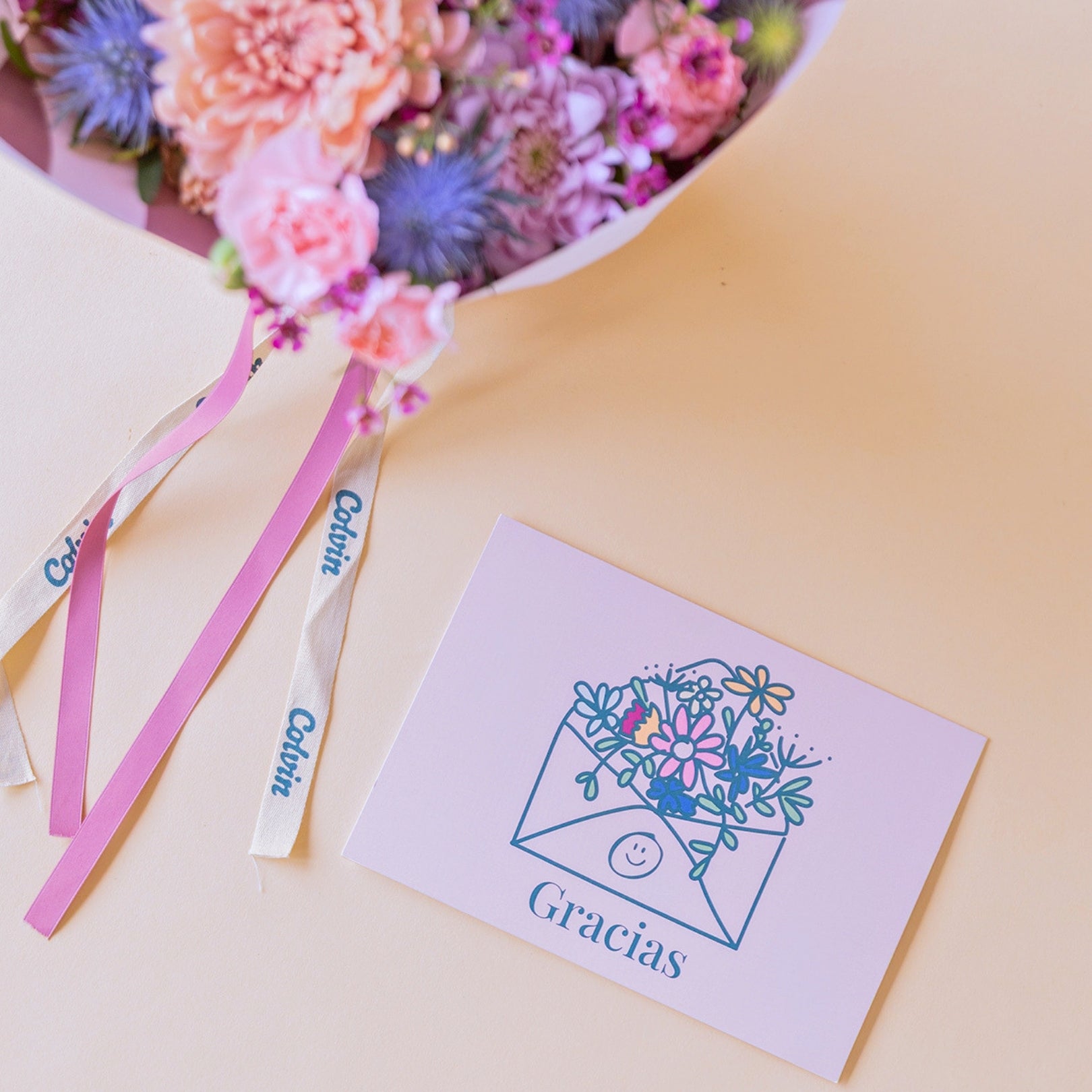 Primer plano de la tarjeta de agradecimiento "Gracias" al lado del ramo floral de tonos rosas Guiño sobre fondo crema.