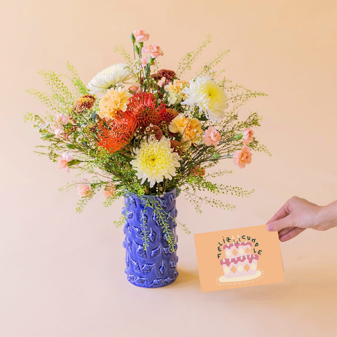 Ramo de flores de verano La Jarana con nutans naranjas y crisantemos en jarrón de cerámica azul texturizado junto a una mano que sostiene una tarjeta de felicitación de Feliz Cumpleaños sobre un fondo color crema.