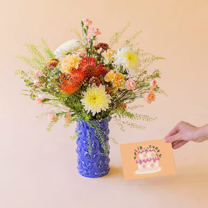 Ramo de flores de verano La Jarana con nutans naranjas y crisantemos en jarrón de cerámica azul texturizado junto a una mano que sostiene una tarjeta de felicitación de Feliz Cumpleaños sobre un fondo color crema.