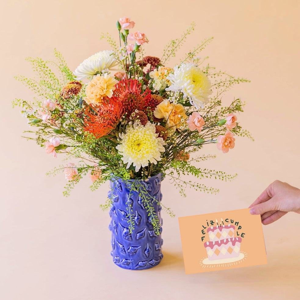 Ramo de flores de verano La Jarana con nutans naranjas y crisantemos en jarrón de cerámica azul texturizado junto a una mano que sostiene una tarjeta de felicitación de Feliz Cumpleaños sobre un fondo color crema.