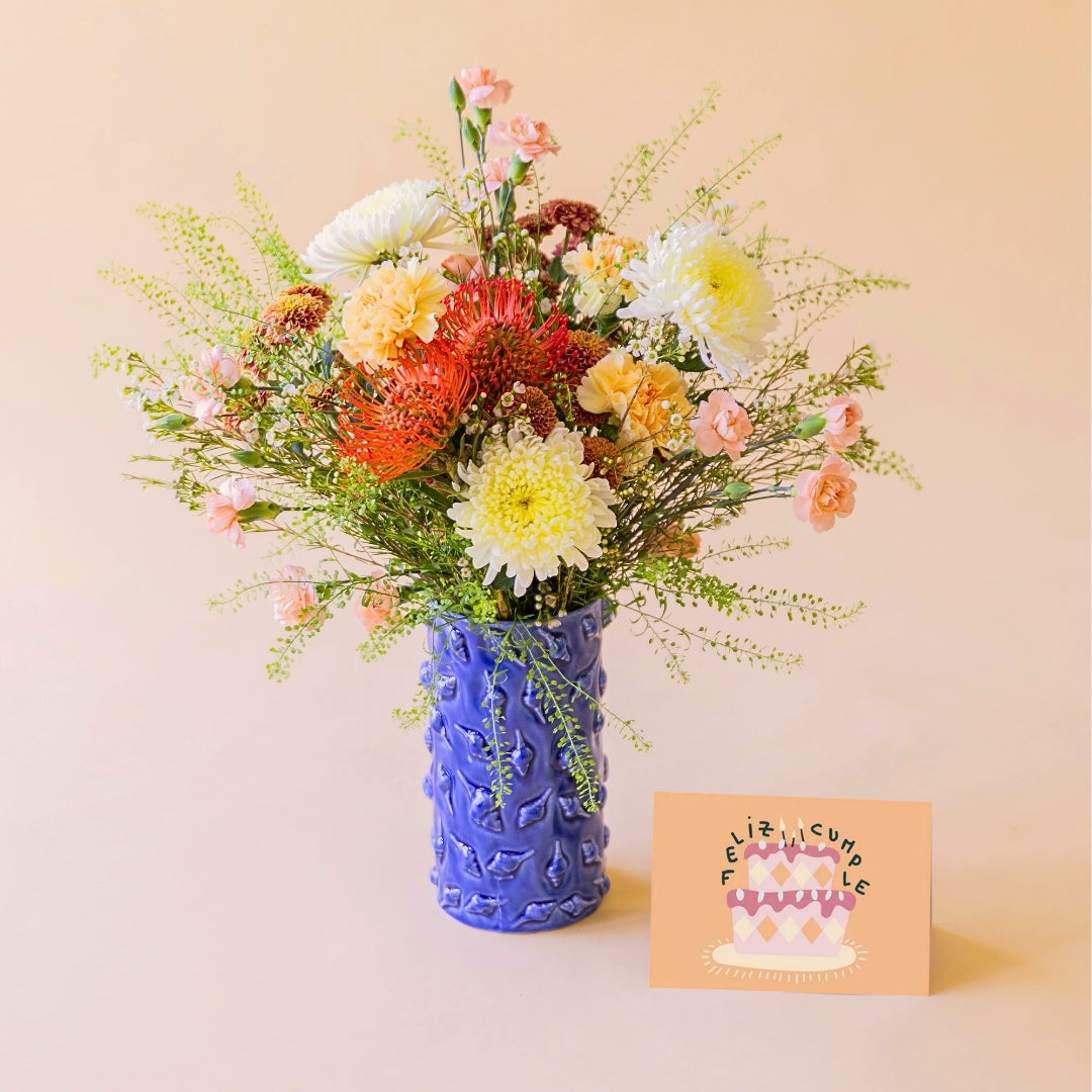Ramo de flores de verano La Jarana con nutans naranjas y crisantemos en jarrón de cerámica azul texturizado con una tarjeta de felicitación de Feliz Cumpleaños.