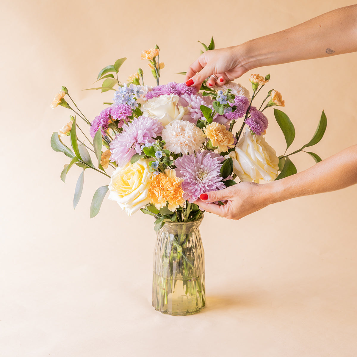 Manos colocando ramo de flores pastel de Colvin con crisantemos lilas, rosas amarillas, claveles melocotón y no me olvides en jarrón de cristal