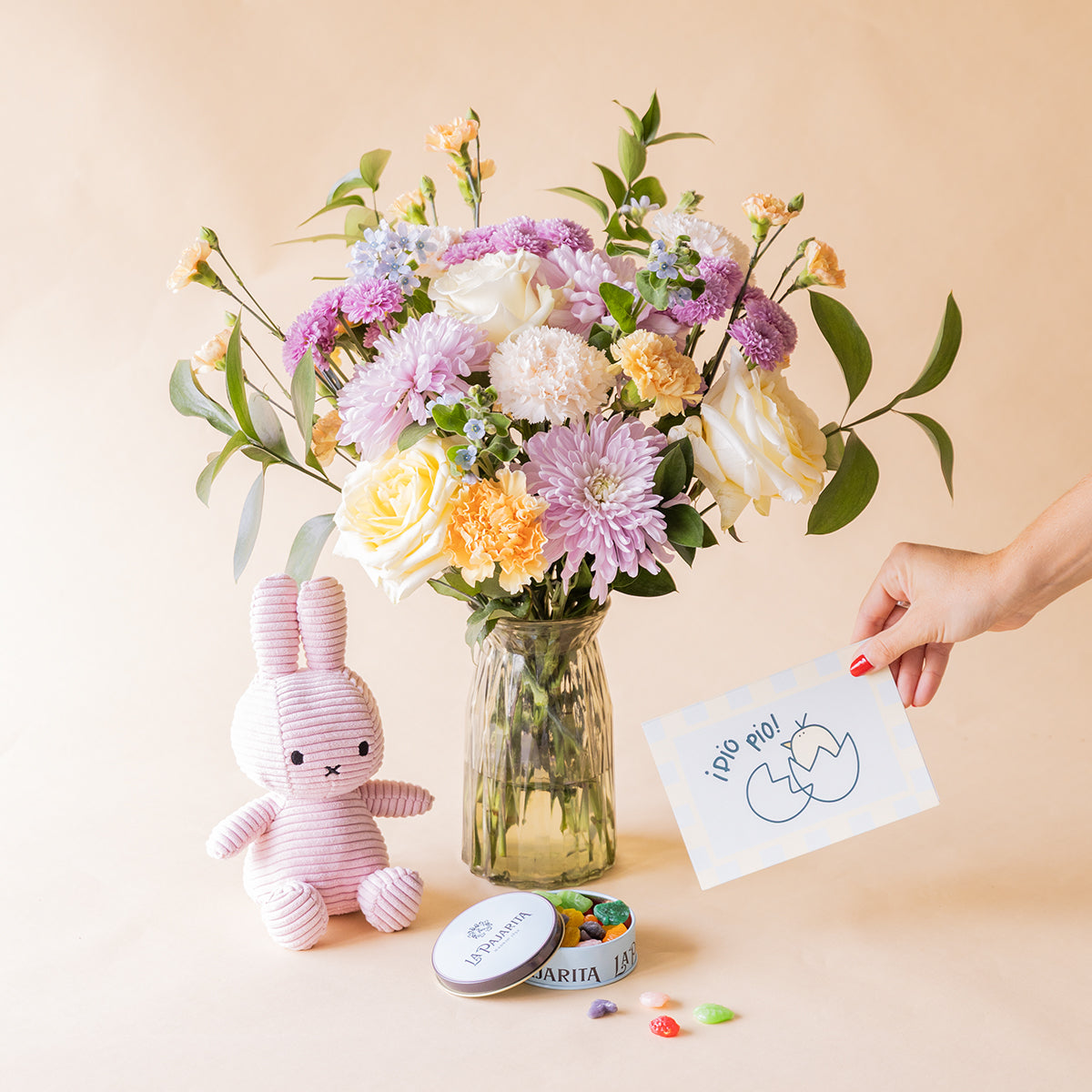 Mano sosteniendo tarjeta "¡Pío Pío!" junto a ramo de flores pastel de Colvin en jarrón de cristal con peluche Miffy rosa y lata de gominolas La Pajarita