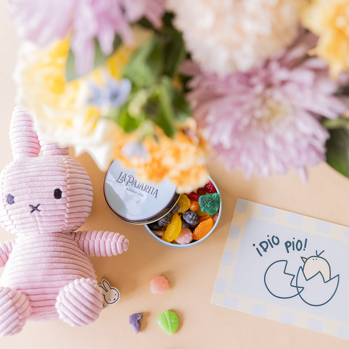 Detalle de peluche Miffy rosa de corduroy, lata de gominolas La Pajarita abierta y tarjeta "¡Pío Pío!" junto a ramo de flores pastel de Colvin desenfocado
