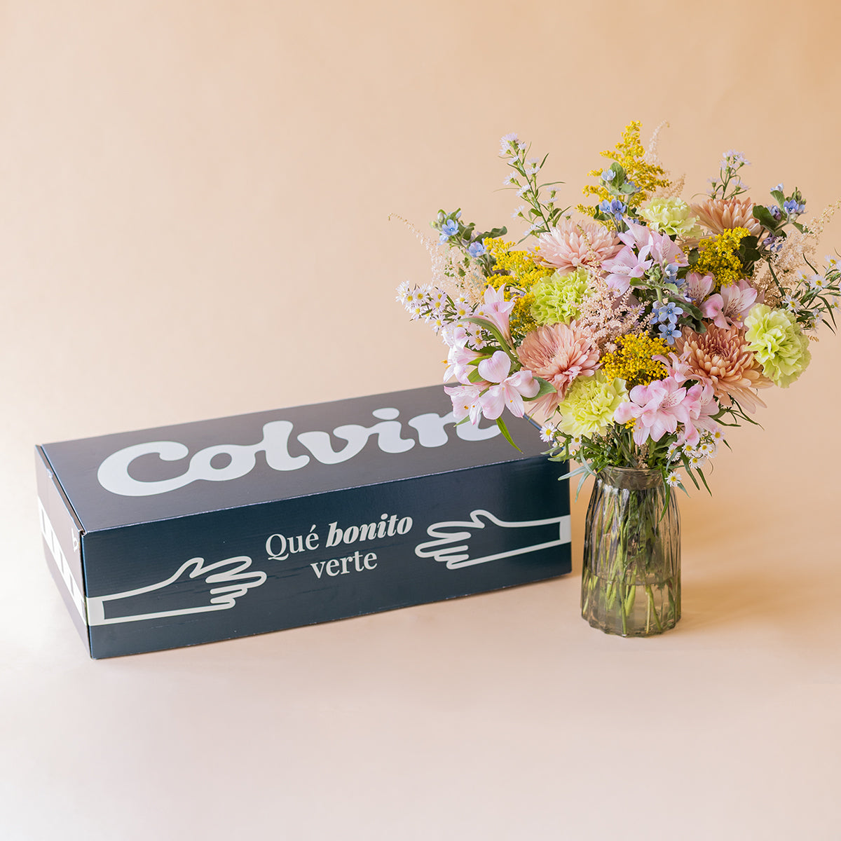 Ramo Colvin silvestre de crisantemos melocotón, claveles verdes y flores silvestres en jarrón de cristal junto a la caja de envío Colvin sobre fondo beige