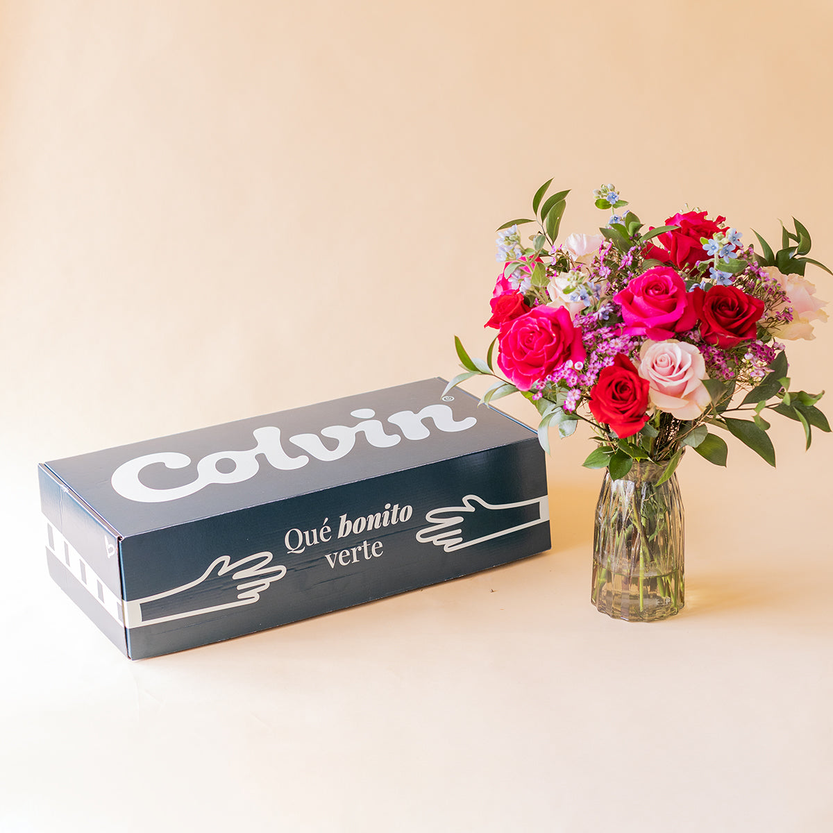 Ramo de rosas fucsias y rojas Colvin El Encanto en jarrón de cristal junto a la caja de envío Colvin sobre fondo beige