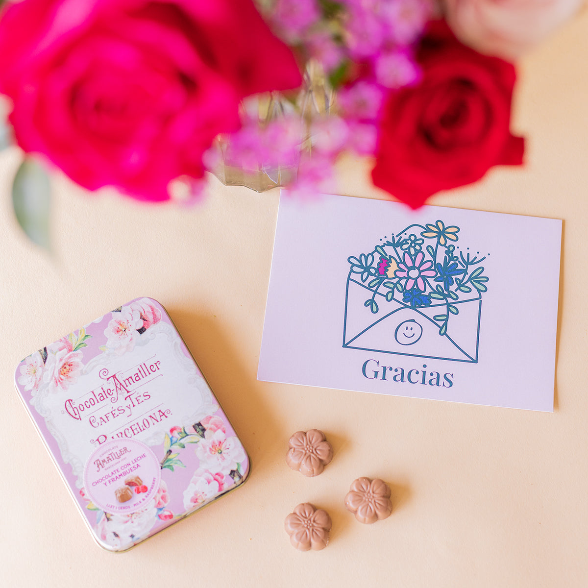 Detalle de lata de bombones florales Amatller y tarjeta Gracias Colvin con rosas fucsias y rojas desenfocadas al fondo