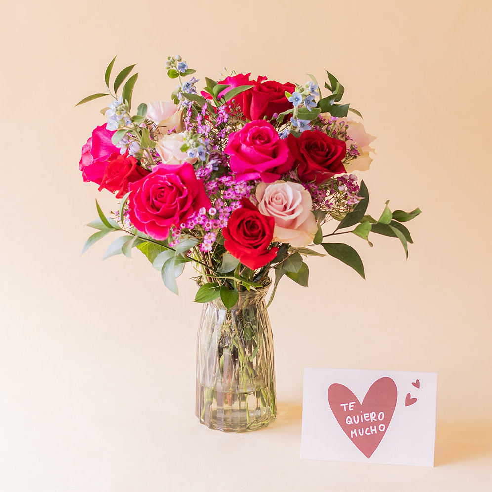 Ramo de rosas fucsias y rojas Colvin El Encanto en jarrón de cristal con tarjeta Te quiero mucho sobre fondo beige