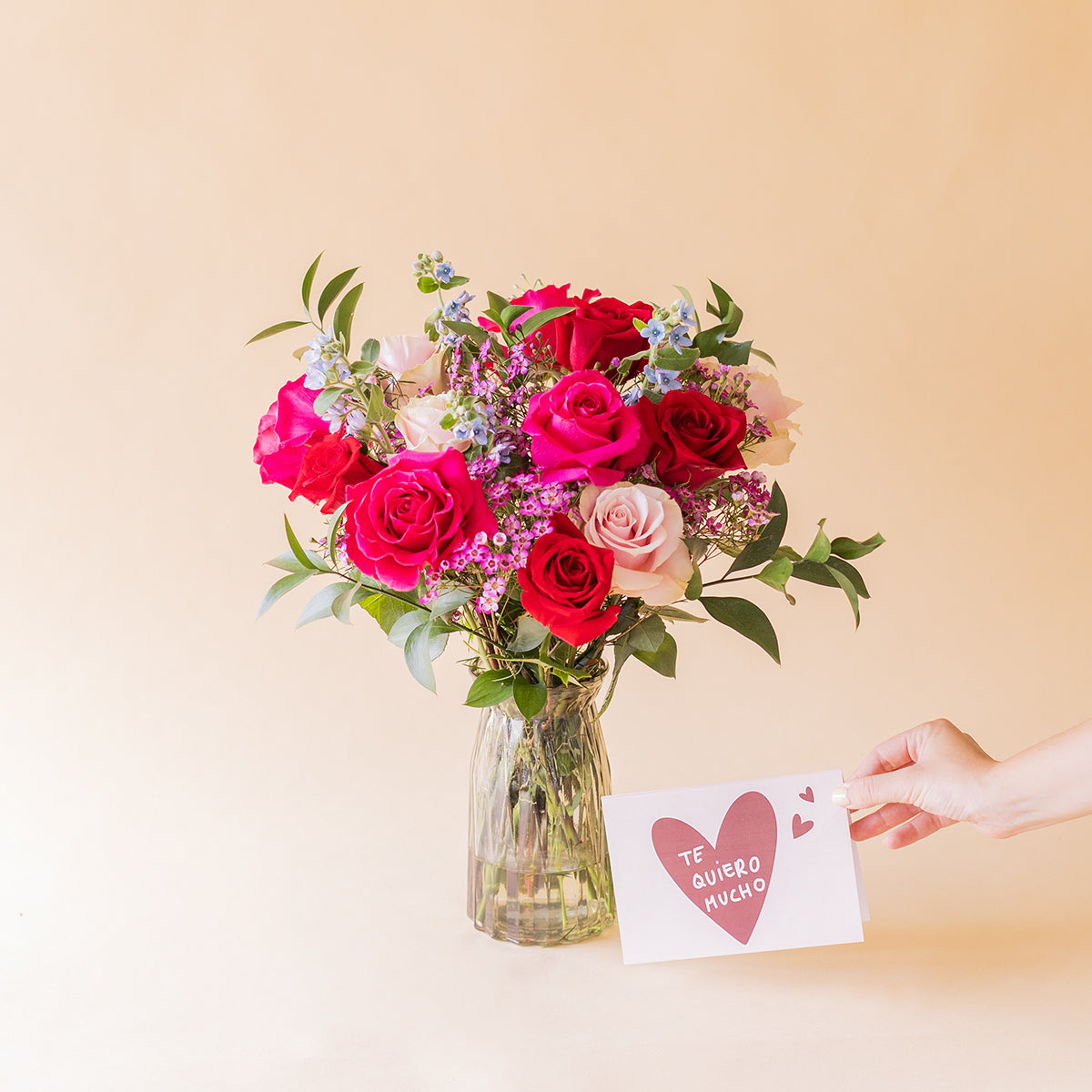 Mano sosteniendo la tarjeta Te quiero mucho junto al ramo de rosas fucsias y rojas Colvin El Encanto en jarrón de cristal