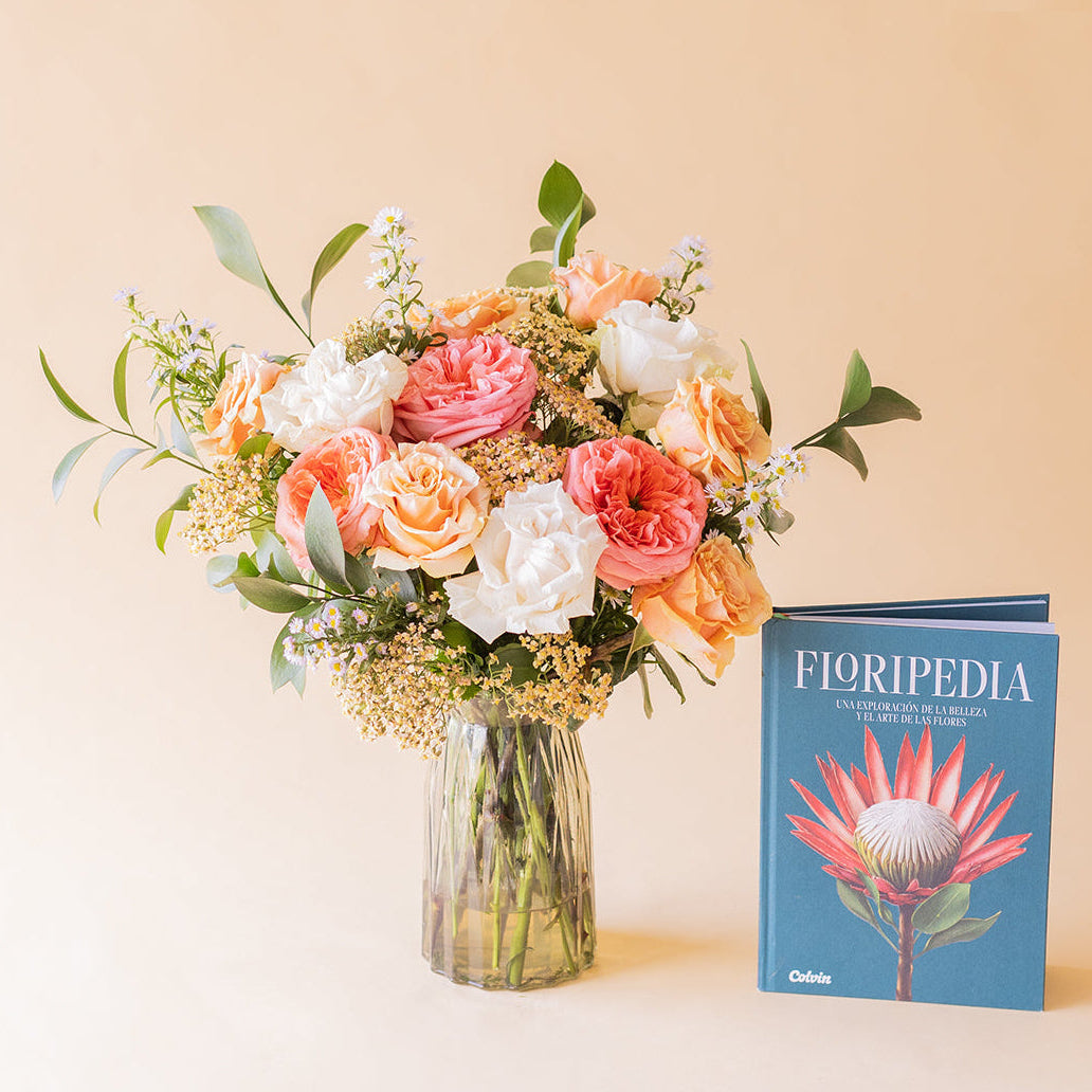 Ramo Colvin romántico de rosas melocotón, rosa coral y blancas en jarrón de cristal junto al libro Floripedia de Colvin sobre fondo beige