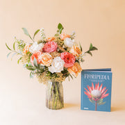 Ramo Colvin romántico de rosas melocotón, rosa coral y blancas en jarrón de cristal junto al libro Floripedia de Colvin sobre fondo beige