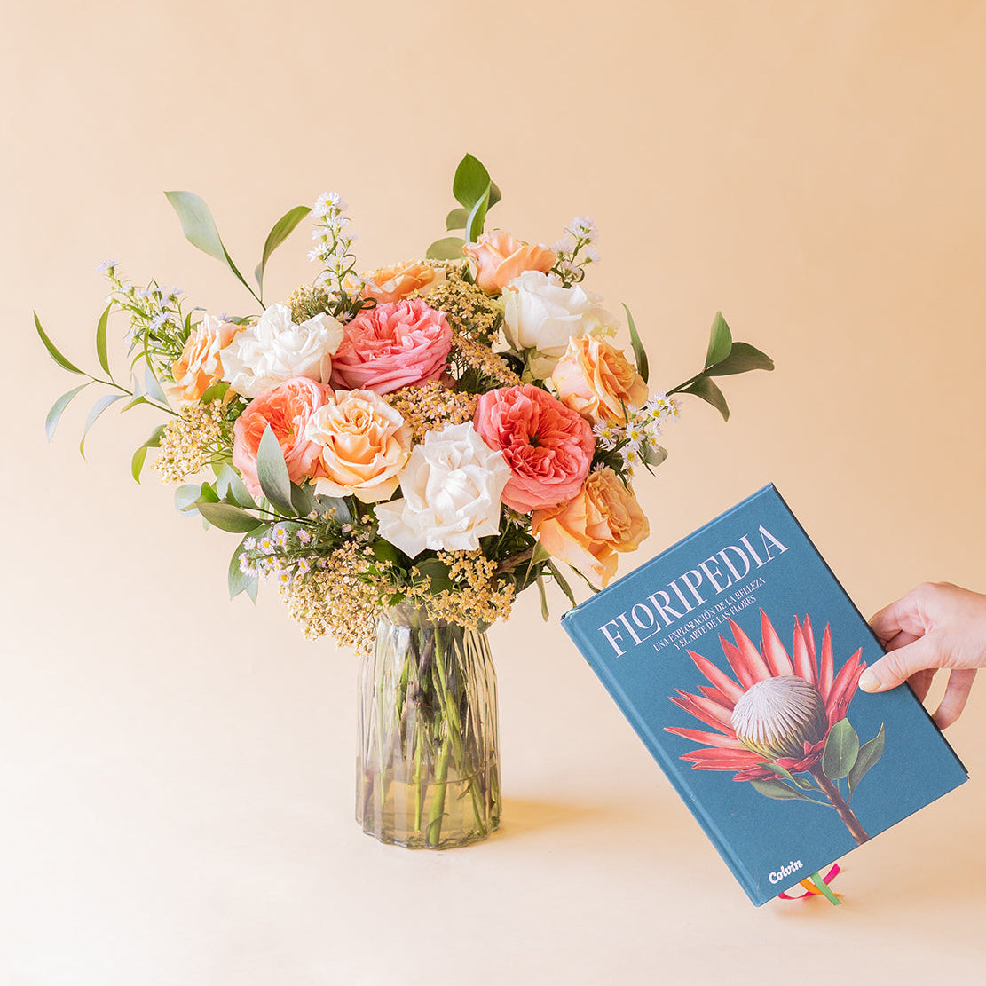 Mano sosteniendo el libro Floripedia de Colvin junto al ramo romántico de rosas melocotón, rosa coral y blancas en jarrón de cristal sobre fondo beige