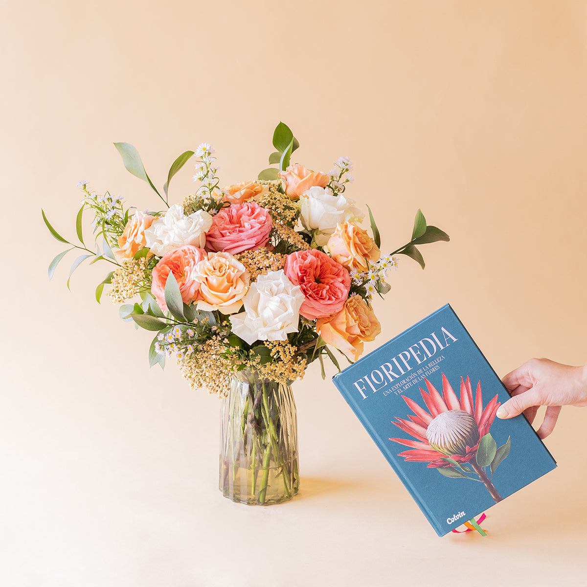 Mano sosteniendo el libro Floripedia de Colvin junto al ramo romántico de rosas melocotón, rosa coral y blancas en jarrón de cristal sobre fondo beige