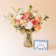 amo Colvin de rosas melocotón, rosa coral y blancas en jarrón de cristal con tarjeta Te lo mereces apoyada sobre fondo beige