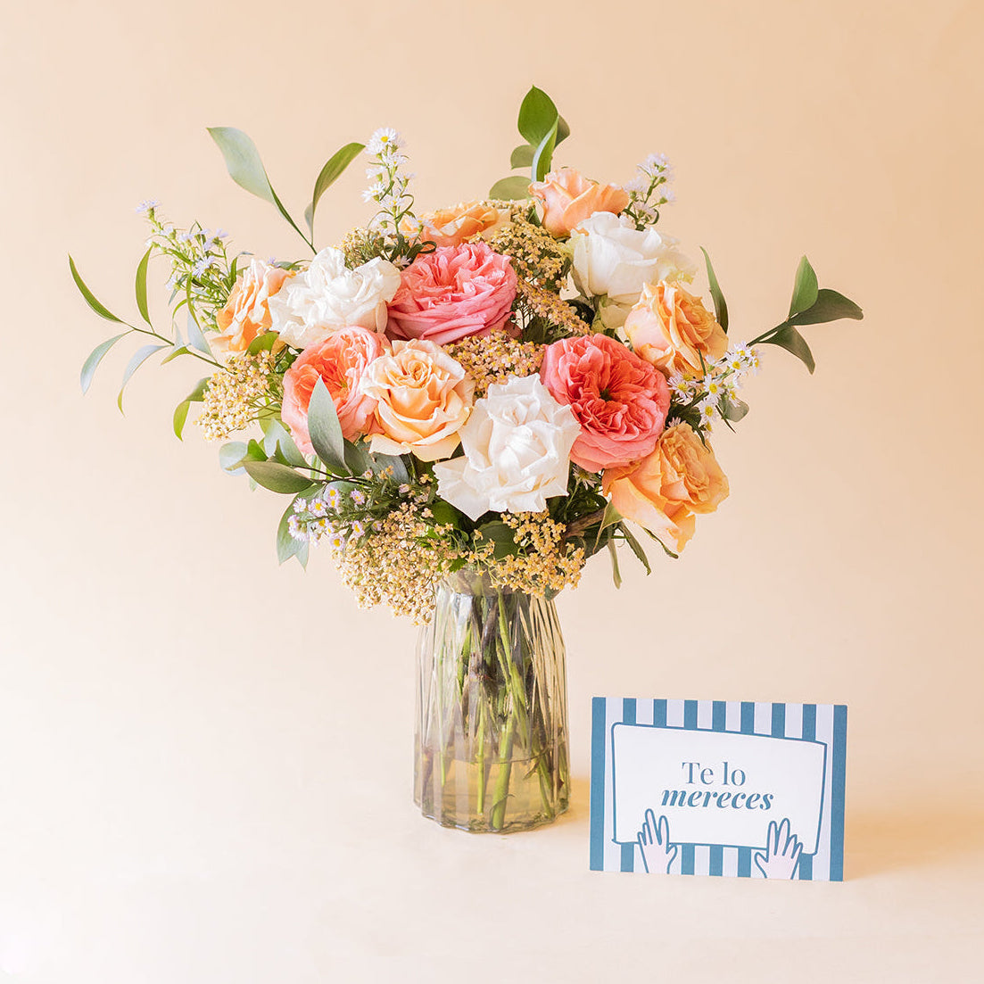 amo Colvin de rosas melocotón, rosa coral y blancas en jarrón de cristal con tarjeta Te lo mereces apoyada sobre fondo beige