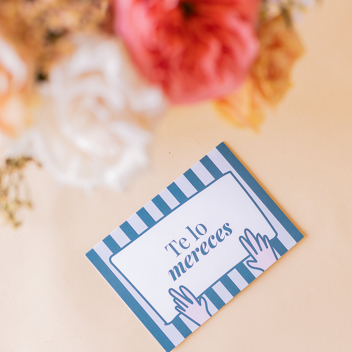 Detalle de la tarjeta Colvin Te lo mereces con rayas azules y rosas melocotón, coral y blancas desenfocadas al fondo