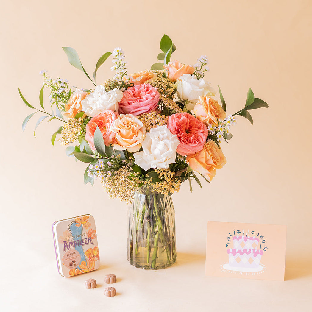 Ramo Colvin de rosas melocotón, rosa y blancas en jarrón de cristal con lata de bombones Amatller capuccino y tarjeta Feliz Cumpleaños sobre fondo beige