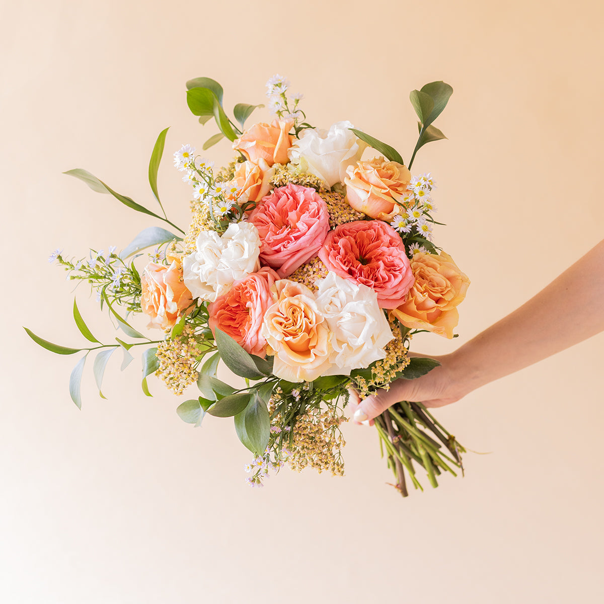 Mano sujetando el ramo Colvin de rosas melocotón, rosa y blancas con flores silvestres y follaje verde sobre fondo beige