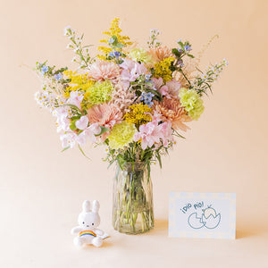 Ramo Colvin silvestre de crisantemos melocotón, claveles verdes, alstroemerias rosas y flores silvestres en jarrón de cristal con llavero Miffy arcoíris y tarjeta ¡Dío Pío! sobre fondo beige