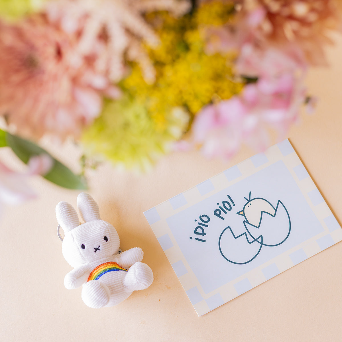 Detalle del llavero Miffy arcoíris y tarjeta ¡Dío Pío! Colvin con crisantemos melocotón y flores silvestres desenfocadas al fondo