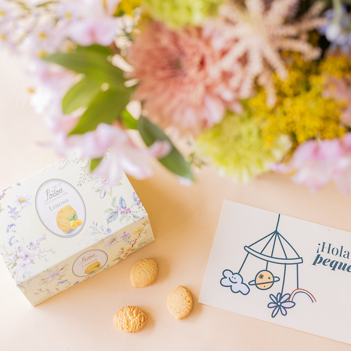 Detalle de la caja de galletas Loison limón y tarjeta ¡Hola peque! Colvin con flores silvestres del ramo desenfocadas al fondo