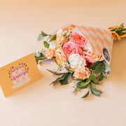 Ramo Colvin de rosas melocotón, rosa y blancas con envoltorio de rombos y tarjeta Feliz Cumpleaños sobre fondo beige