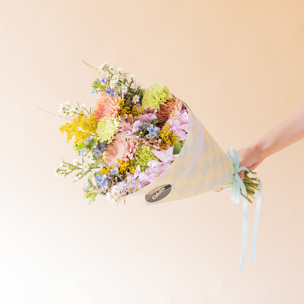Mano sujetando el ramo Colvin silvestre con envoltorio de cuadros azules y lazo celeste, con crisantemos melocotón, hortensias rosas y flores silvestres sobre fondo beige