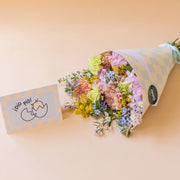 Ramo Colvin silvestre de crisantemos melocotón, hortensias rosas y flores silvestres con envoltorio de cuadros azules y tarjeta ¡Dió Pío! sobre fondo beige