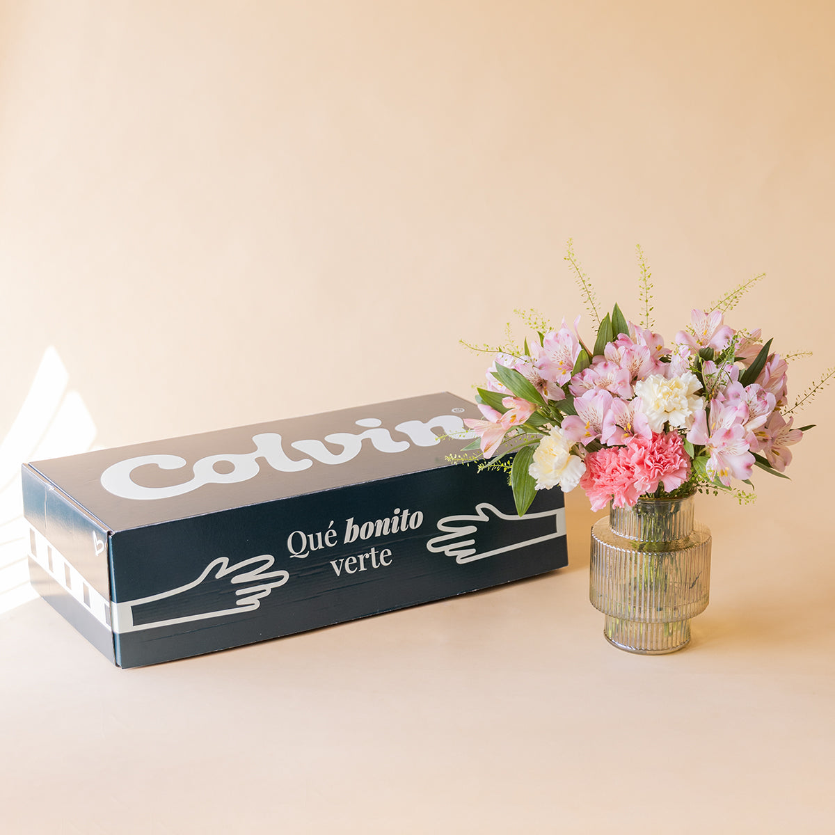 Ramo de alstroemerias rosas, claveles blancos y rosas de Colvin en jarrón de cristal acanalado junto a caja de envío Colvin con texto Qué bonito verte, sobre fondo melocotón