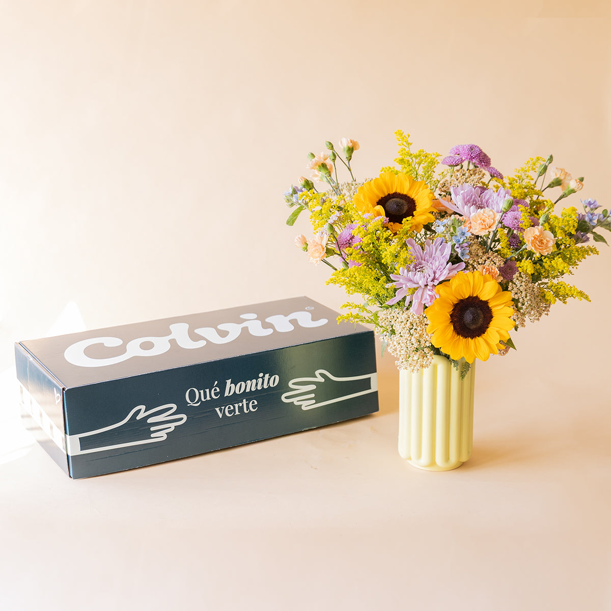 Ramo de girasoles y crisantemos lilas en jarrón acanalado amarillo junto a caja de envío Colvin con texto Qué bonito verte, sobre fondo beige