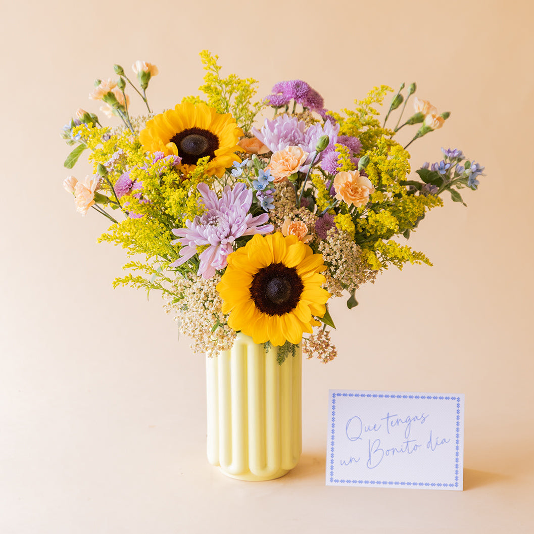 Ramo de girasoles, crisantemos lilas y flores silvestres de Colvin en jarrón acanalado amarillo claro con tarjeta Que tengas un Bonito día, sobre fondo beige