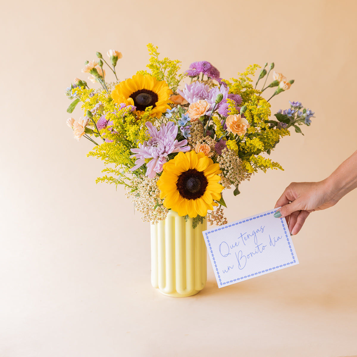 Mano sosteniendo tarjeta Que tengas un Bonito día junto al ramo de girasoles y crisantemos en jarrón amarillo de Colvin, sobre fondo beige
