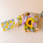 amo de girasoles y crisantemos lilas de Colvin con envoltorio amarillo estampado y tarjeta ilustrada con limones, vista cenital sobre fondo beige