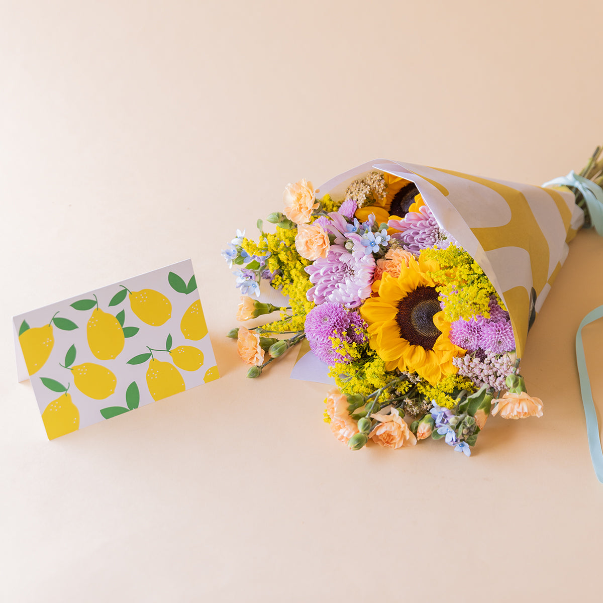 amo de girasoles y crisantemos lilas de Colvin con envoltorio amarillo estampado y tarjeta ilustrada con limones, vista cenital sobre fondo beige