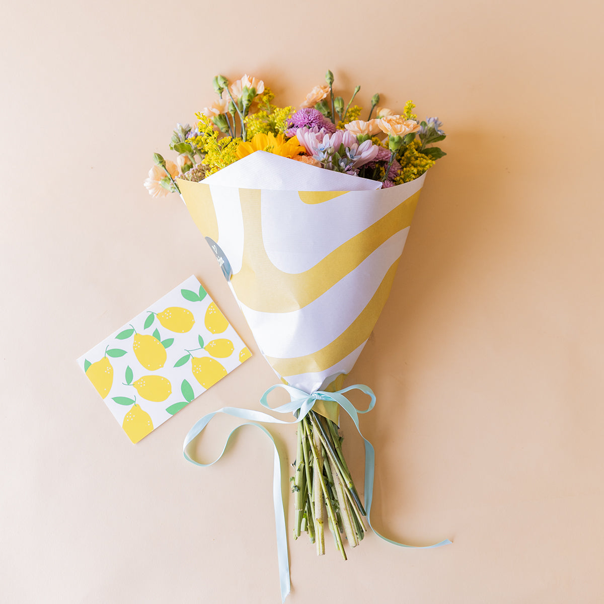 Ramo de girasoles y flores silvestres de Colvin envuelto en papel amarillo estampado, atado con cinta azul, junto a tarjeta de limones sobre fondo beige