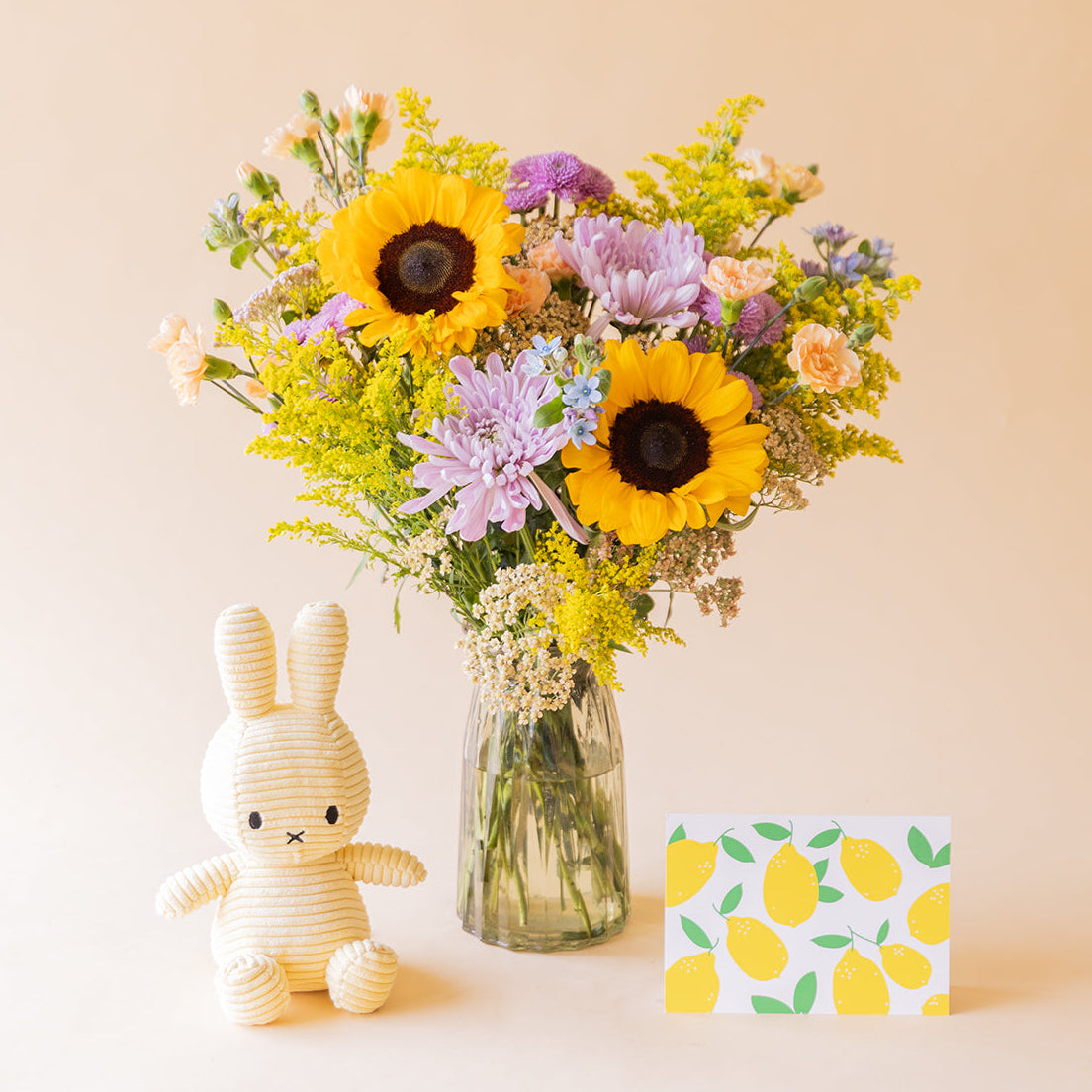 amo de girasoles y crisantemos lilas de Colvin con peluche Miffy y tarjeta de limones, vista frontal sobre fondo beige