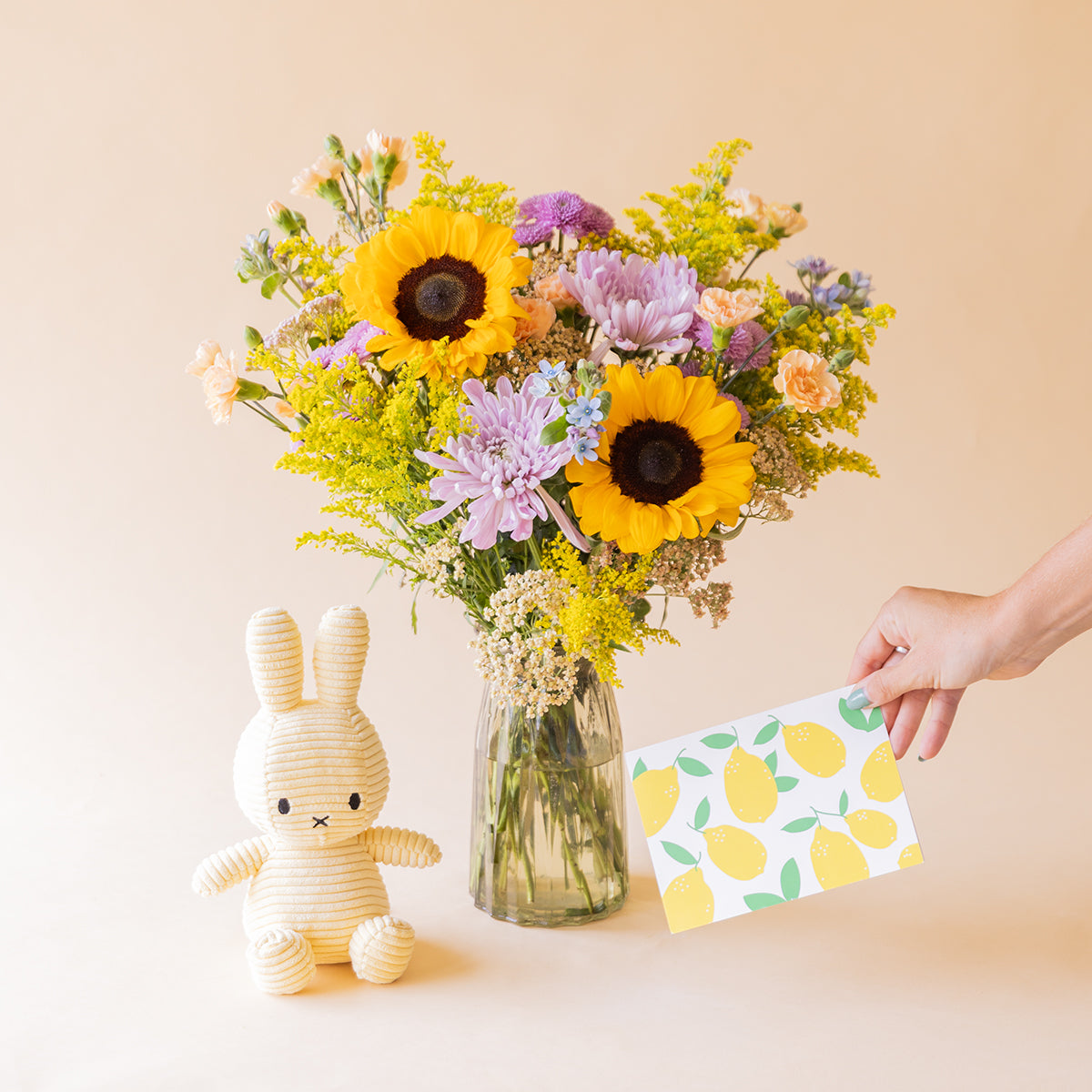 Ramo de girasoles y crisantemos en jarrón de cristal junto a peluche Miffy y mano sosteniendo tarjeta de limones, Colvin