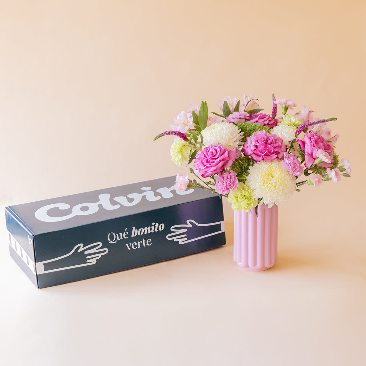 Ramo de rosas fucsias y flores rosas de Colvin en jarrón acanalado rosa junto a caja de envío Colvin con texto Qué bonito verte, sobre fondo melocotón