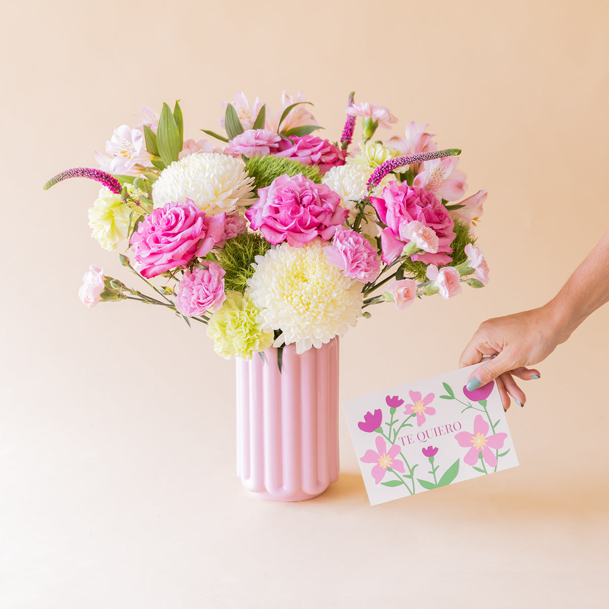 Ramo de rosas fucsias, crisantemos blancos, claveles rosas y alstroemeria de Colvin en jarrón acanalado rosa, con mano sosteniendo tarjeta Te Quiero, sobre fondo melocotón
