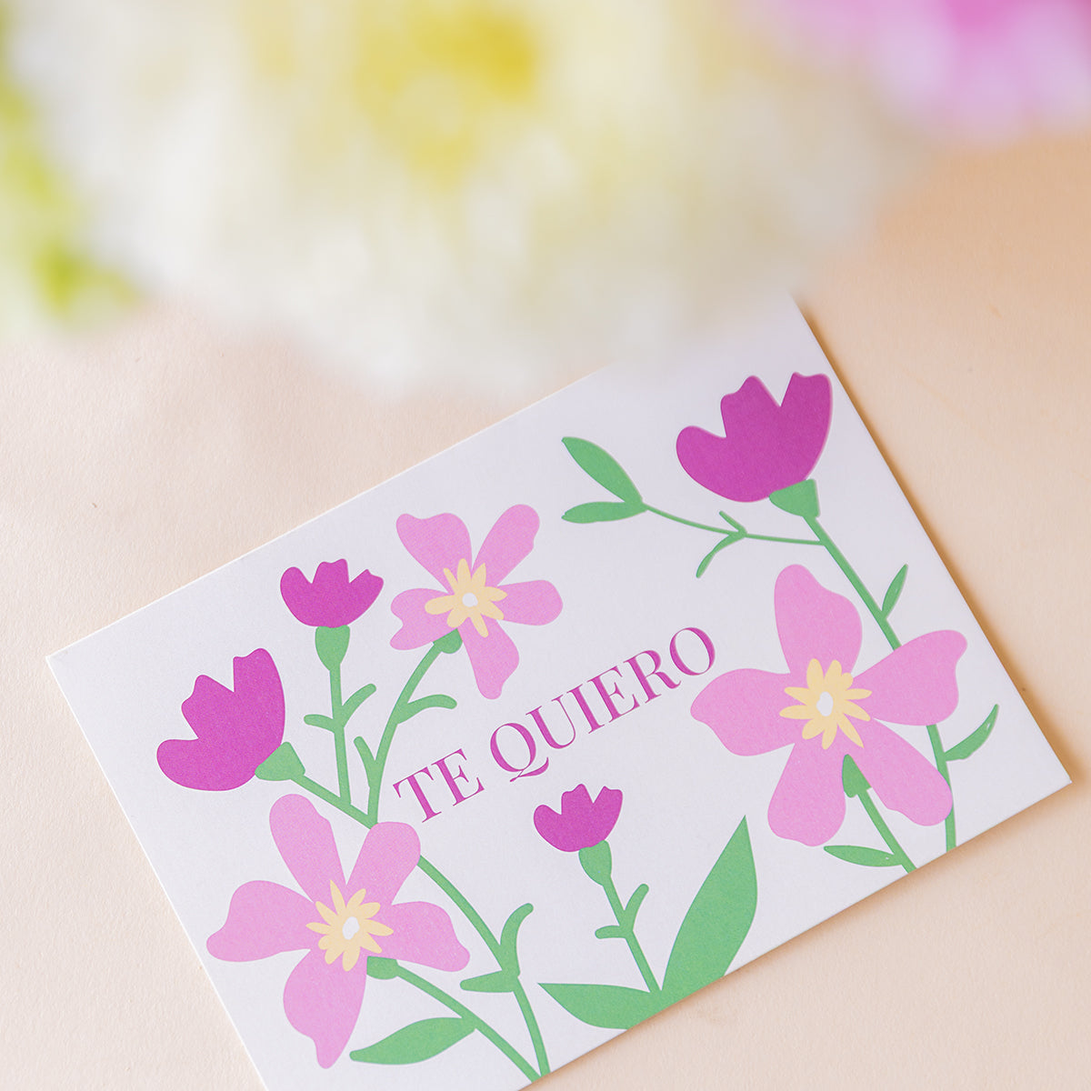 Tarjeta Te Quiero ilustrada con flores rosas y moradas sobre fondo blanco, incluida en el ramo de rosas fucsias de Colvin
