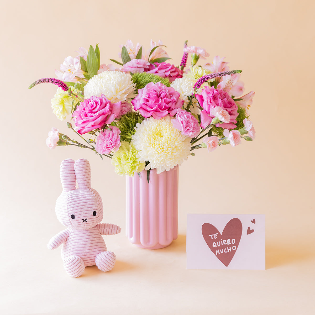 Ramo de rosas fucsias, crisantemos blancos, alstroemeria rosa y veronica lila de Colvin en jarrón acanalado rosa con peluche Miffy lila y tarjeta Te quiero mucho, sobre fondo melocotón